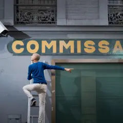 Un homme sur une échelle pointe du doigt vers une caméra de surveillance devant un commissariat, prêt à dénoncer quelqu'un à la préfecture.