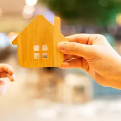 Une main tient une maison en bois, une autre main tendue vers elle.