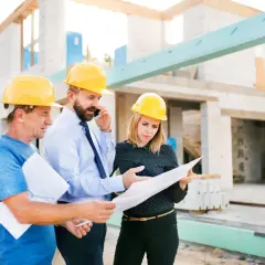 Trois personnes casquées discutent des plans sur un chantier. L'un d'eux est au téléphone, peut-être pour résoudre un litige travaux.