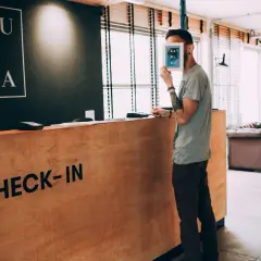 Un homme cache son visage derrière une carte, à la réception d'un hôtel. Le panneau "CHECK-IN" est visible. Un cas de surbooking hotel ?