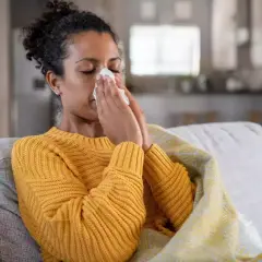 Femme malade sur un canapé, se mouchant dans un mouchoir. Elle est couverte d'une couverture. Cela pourrait être un arrêt maladie pendant préavis démission.
