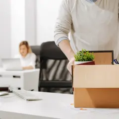 Un homme emporte sa boîte de démission, contenant des fournitures de bureau et une plante. Une collègue travaille à l'arrière-plan.
