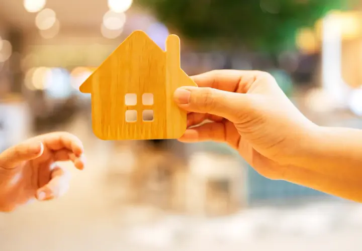 Une main tient une maison en bois, une autre main tendue vers elle.