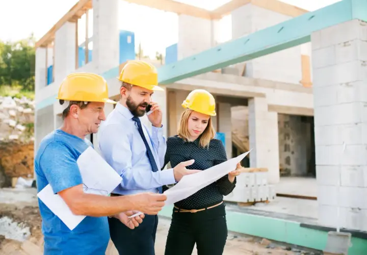 Trois personnes casquées discutent des plans sur un chantier. L'un d'eux est au téléphone, peut-être pour résoudre un litige travaux.