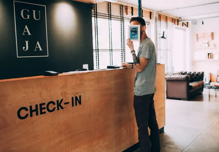 Un homme cache son visage derrière une carte, à la réception d'un hôtel. Le panneau "CHECK-IN" est visible. Un cas de surbooking hotel ?
