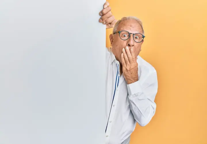 Homme âgé surpris, main sur la bouche, derrière un panneau blanc.