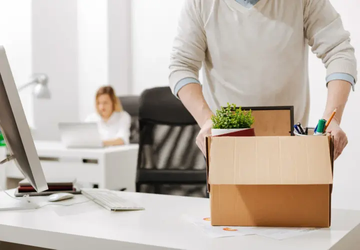 Un homme emporte sa boîte de démission, contenant des fournitures de bureau et une plante. Une collègue travaille à l'arrière-plan.