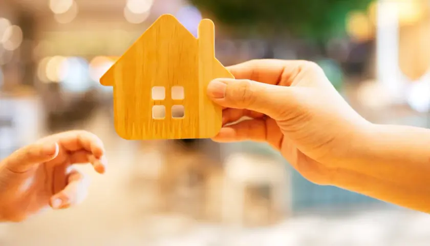 Une main tient une maison en bois, une autre main tendue vers elle.
