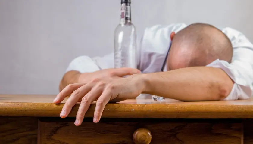 Un homme endormi sur son bureau, une bouteille vide à côté. Les conséquences de l'alcool au travail sont visibles.