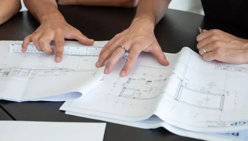 Des mains travaillent sur des plans d'architecte, une règle à la main. Comprendre ces détails est essentiel pour savoir pourquoi faire un devis précis.