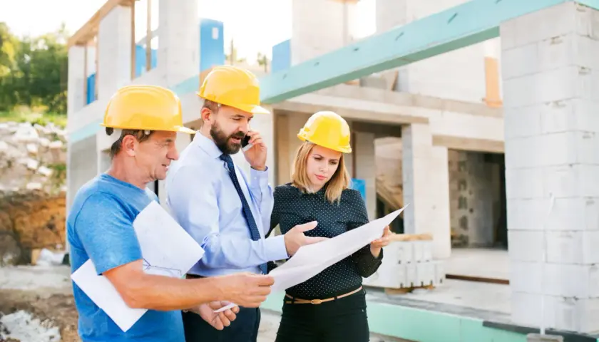 Trois personnes casquées discutent des plans sur un chantier. L'un d'eux est au téléphone, peut-être pour résoudre un litige travaux.