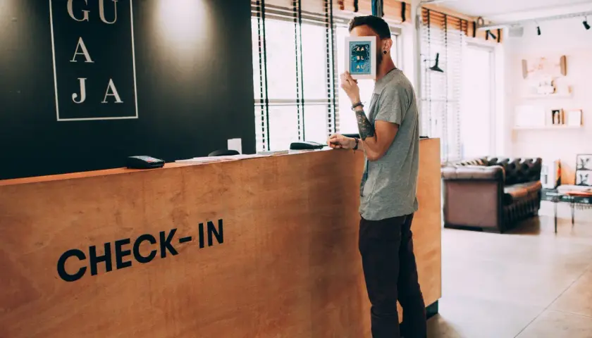 Un homme cache son visage derrière une carte, à la réception d'un hôtel. Le panneau "CHECK-IN" est visible. Un cas de surbooking hotel ?