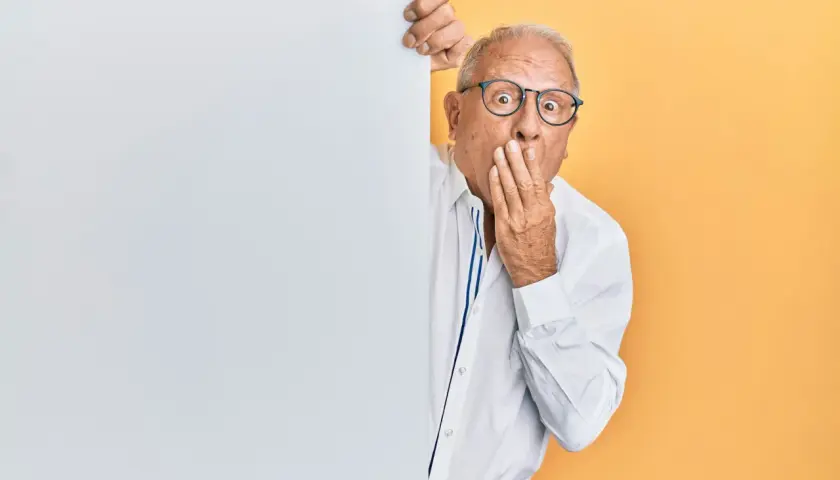 Homme âgé surpris, main sur la bouche, derrière un panneau blanc.