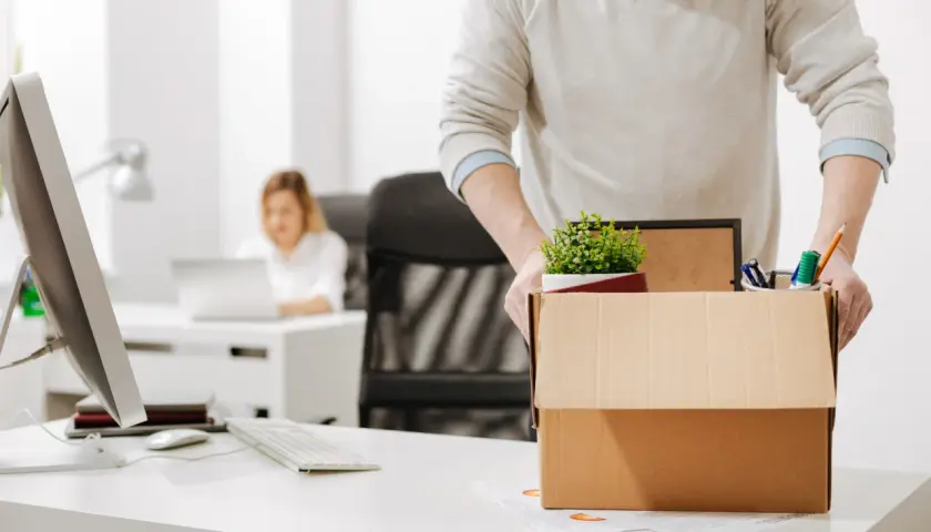 Un homme emporte sa boîte de démission, contenant des fournitures de bureau et une plante. Une collègue travaille à l'arrière-plan.