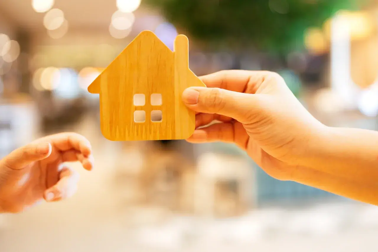 Une main tient une maison en bois, une autre main tendue vers elle.