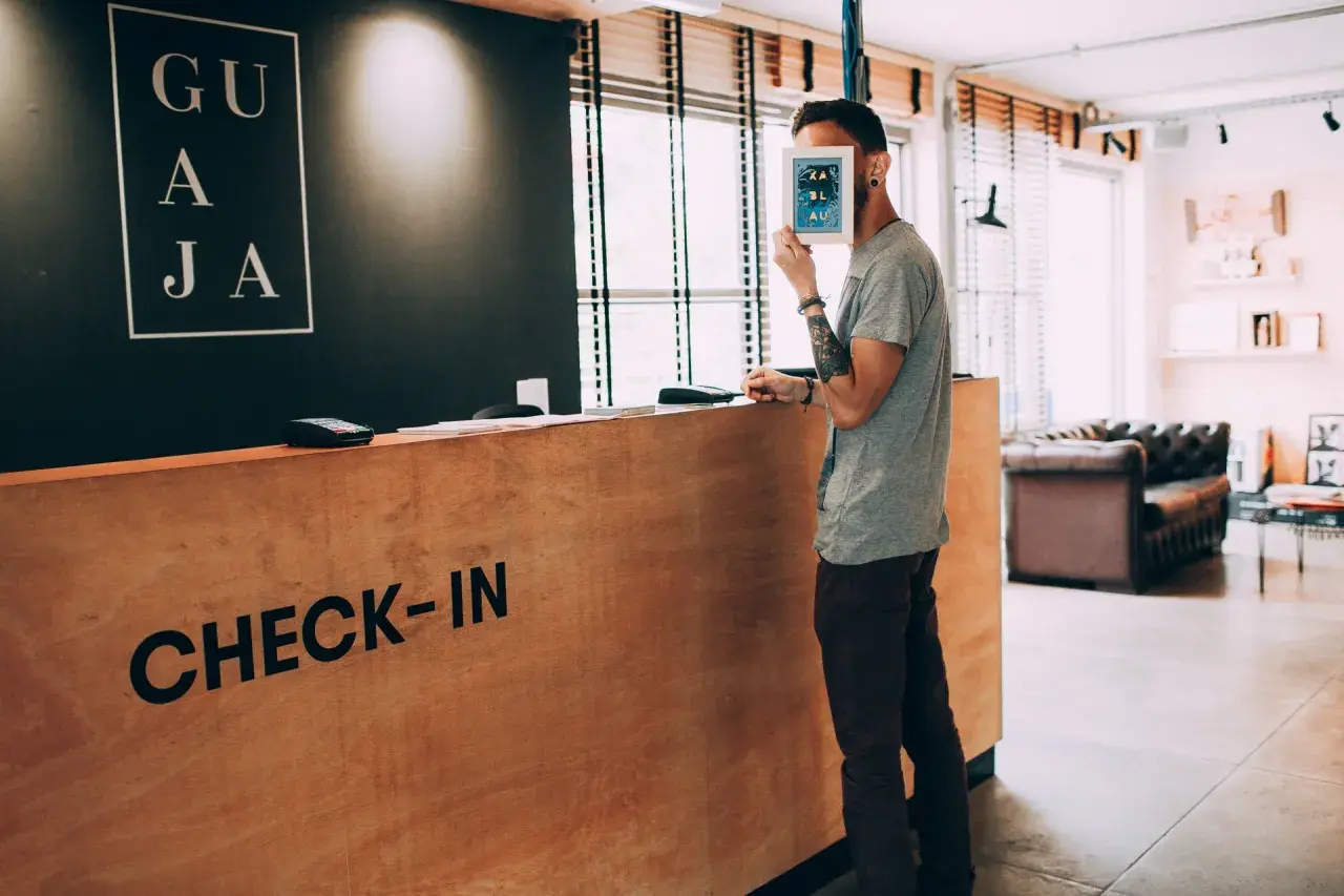 Un homme cache son visage derrière une carte, à la réception d'un hôtel. Le panneau "CHECK-IN" est visible. Un cas de surbooking hotel ?