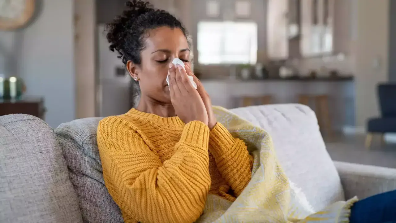 Femme malade sur un canapé, se mouchant dans un mouchoir. Elle est couverte d'une couverture. Cela pourrait être un arrêt maladie pendant préavis démission.