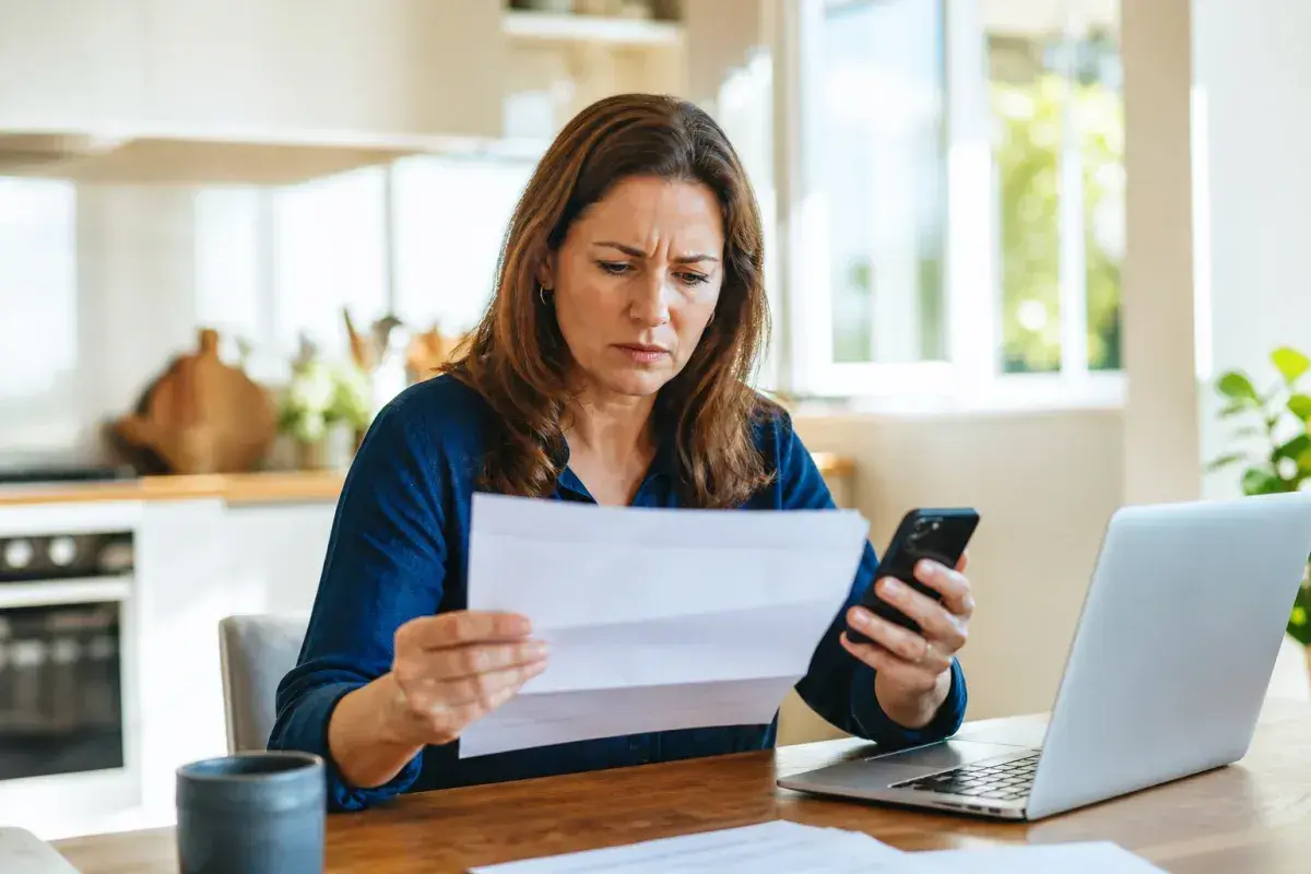 Femme soucieuse regardant une facture et son téléphone, peut-être pour vérifier un prélèvement automatique. Un ordinateur portable est ouvert à côté.