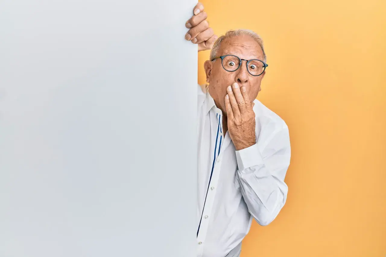 Homme âgé surpris, main sur la bouche, derrière un panneau blanc.