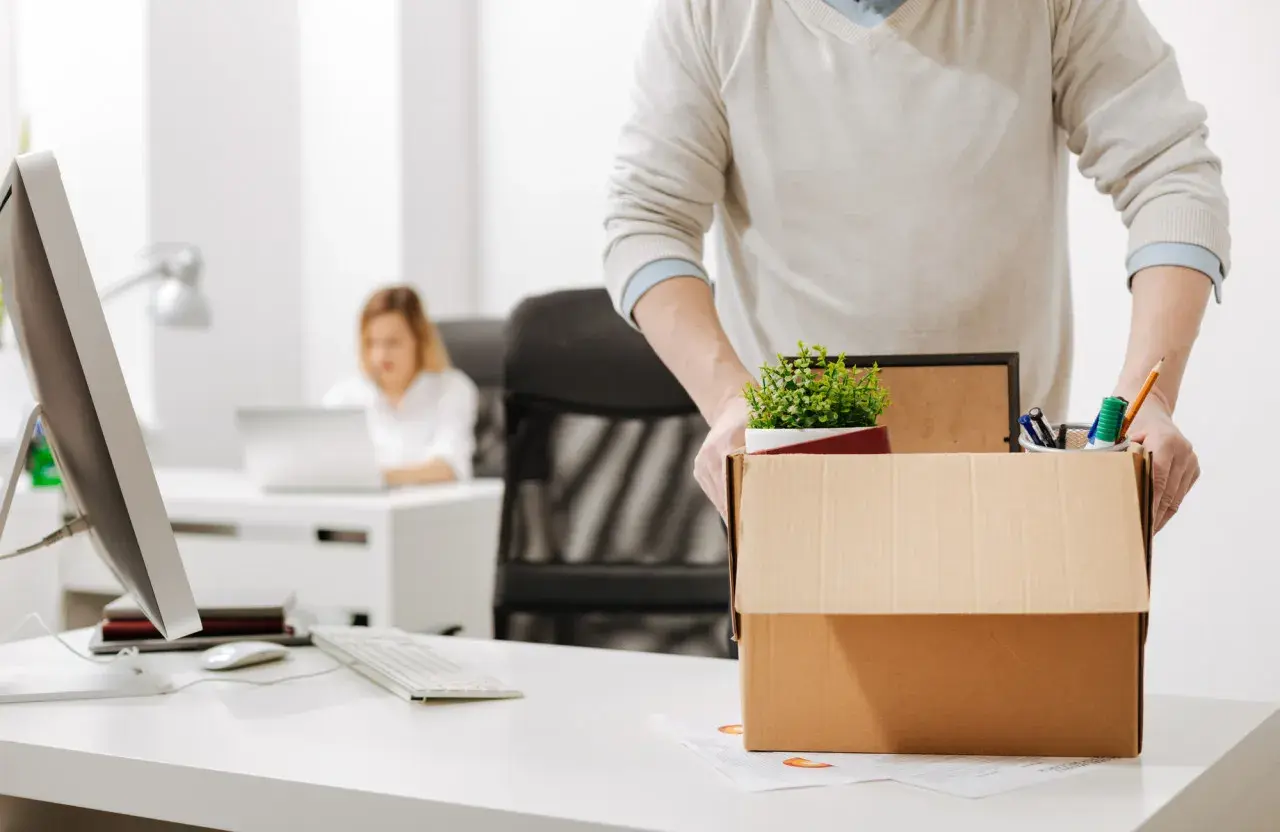 Un homme emporte sa boîte de démission, contenant des fournitures de bureau et une plante. Une collègue travaille à l'arrière-plan.