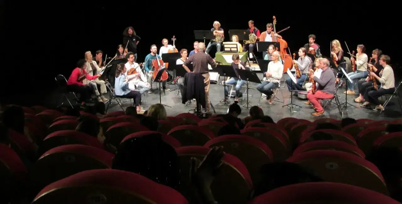 Un orchestre joue sur sc&egrave;ne, dirig&eacute; par un chef. Les musiciens, dont certains jouent du violon, sont assis sur des chaises. L'ambiance rappelle une r&eacute;p&eacute;tition pour la convention collective idcc 1285.