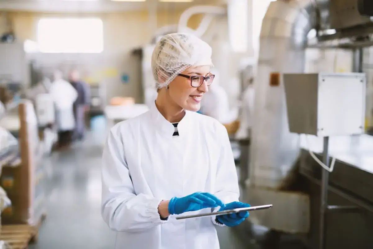 Employ&eacute;e en blouse blanche et charlotte, effectuant un contr&ocirc;le d'hygi&egrave;ne sur tablette dans une usine.